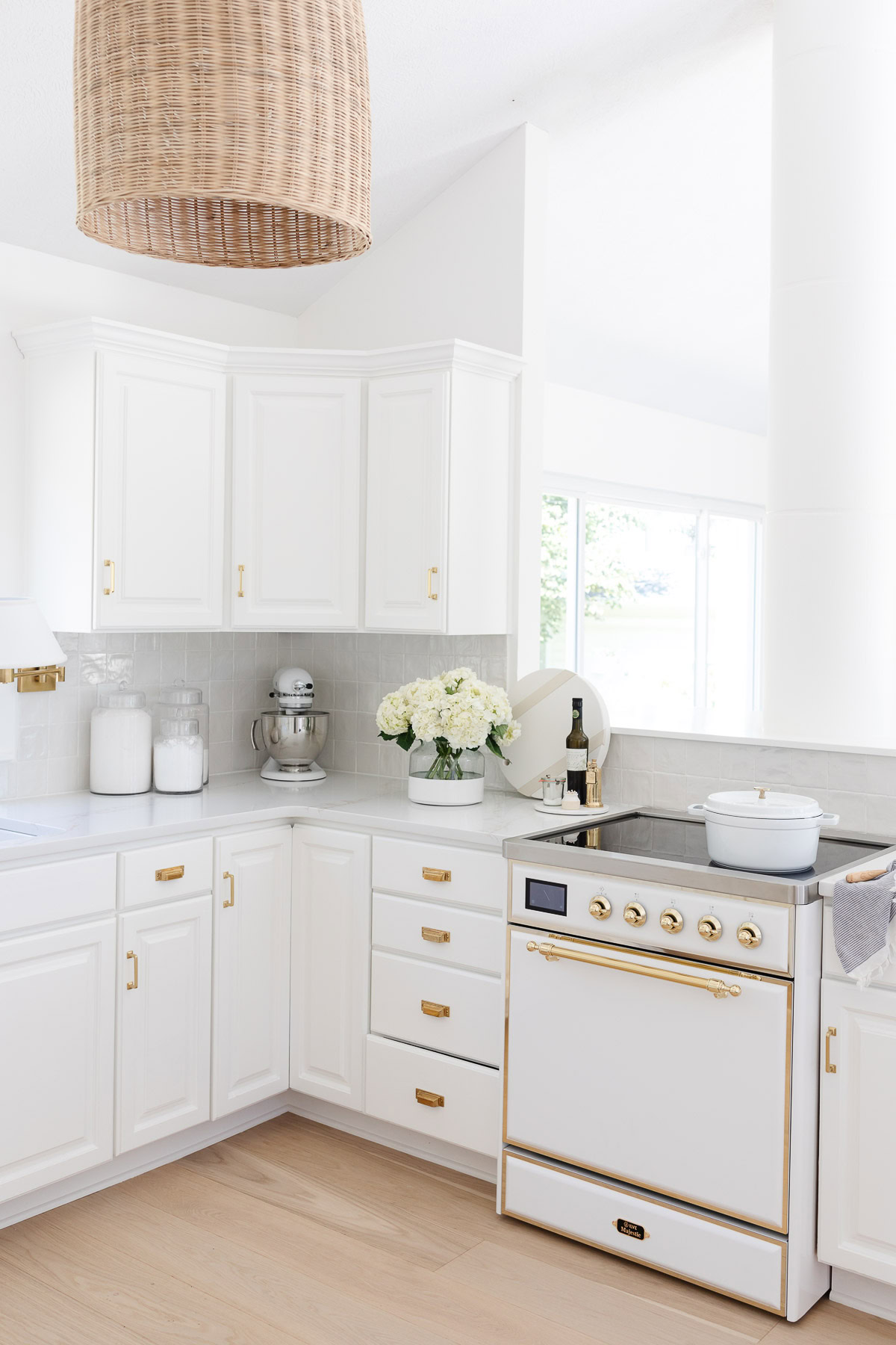 Bright white kitchen with gold hardware, a vintage-style stove, white canisters, a mixer, a vase of flowers, and a woven pendant light atop elegant white oak floors.