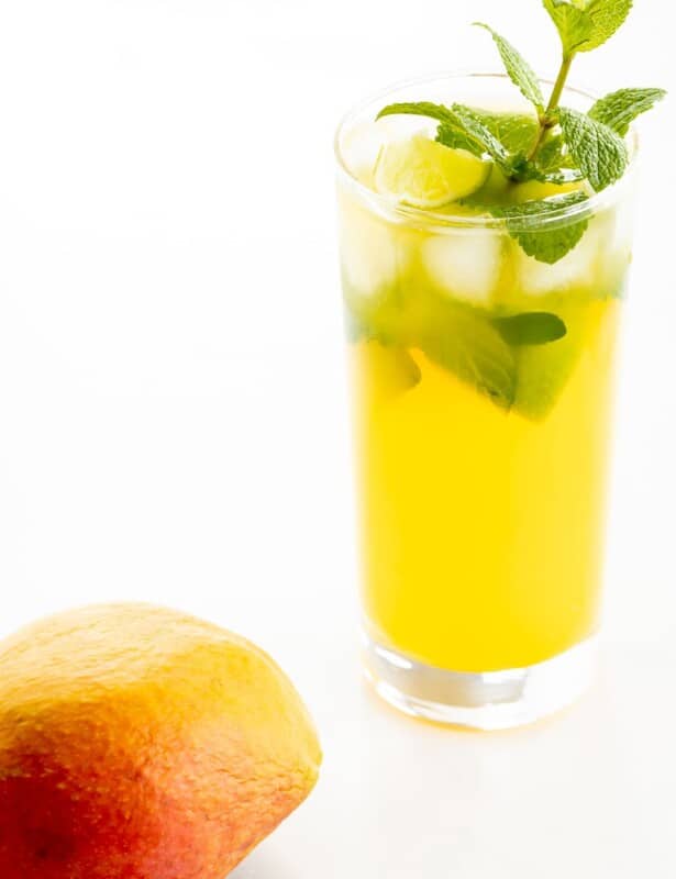 A mango mojito recipe in a clear glass, garnished with a sprig of mint, mango in foreground.