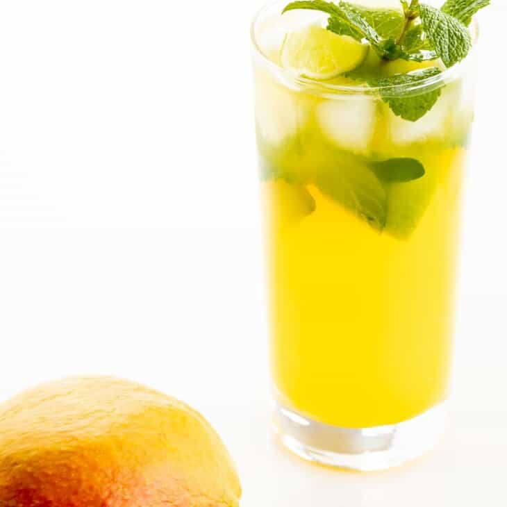 A mango mojito recipe in a clear glass, garnished with a sprig of mint, mango in foreground.