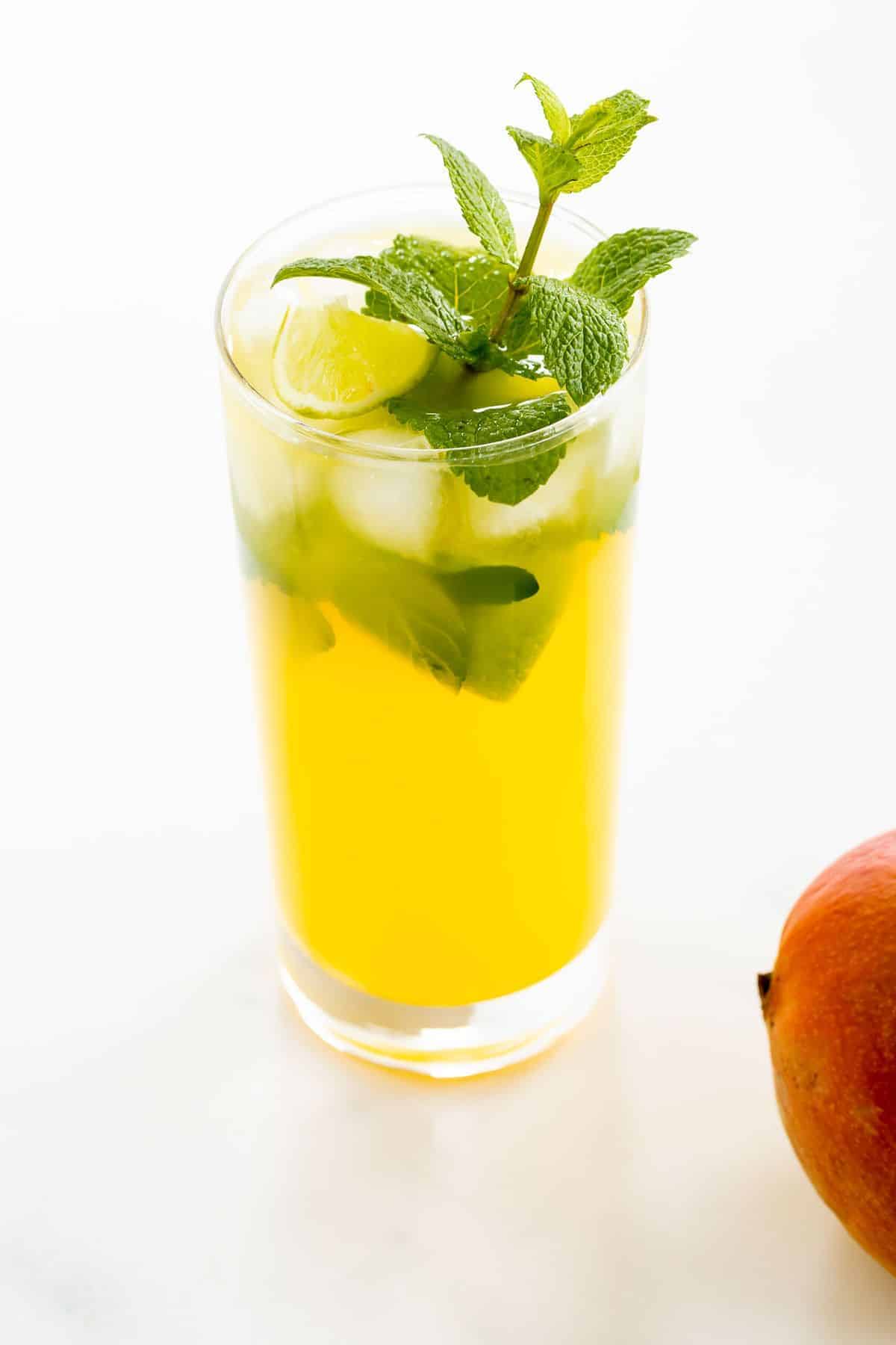 A mango mojito recipe in a clear glass, garnished with a sprig of mint, mango in foreground.