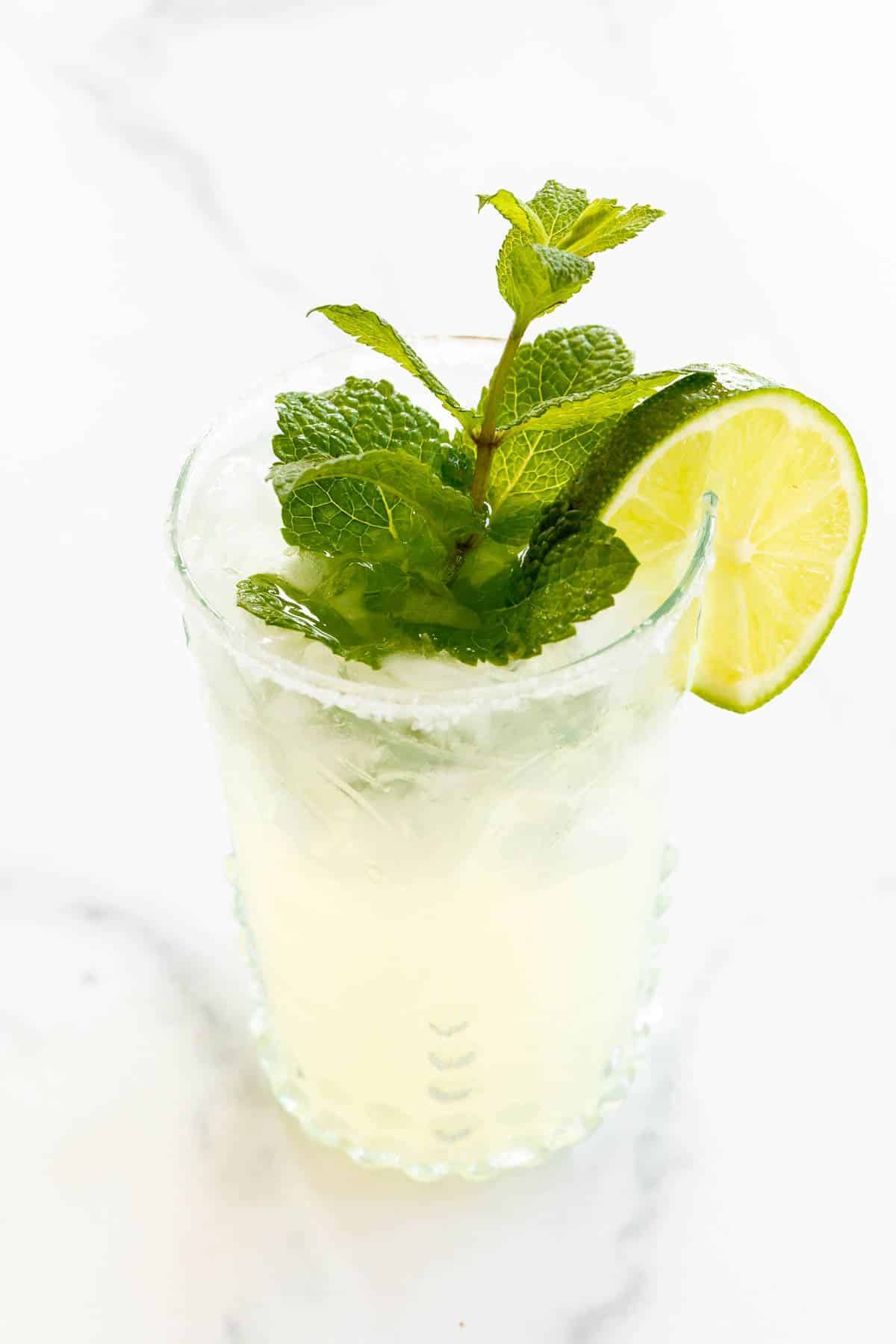 A mint margarita in a clear glass, topped with a sprig of mint and a slice of lime on a white background.