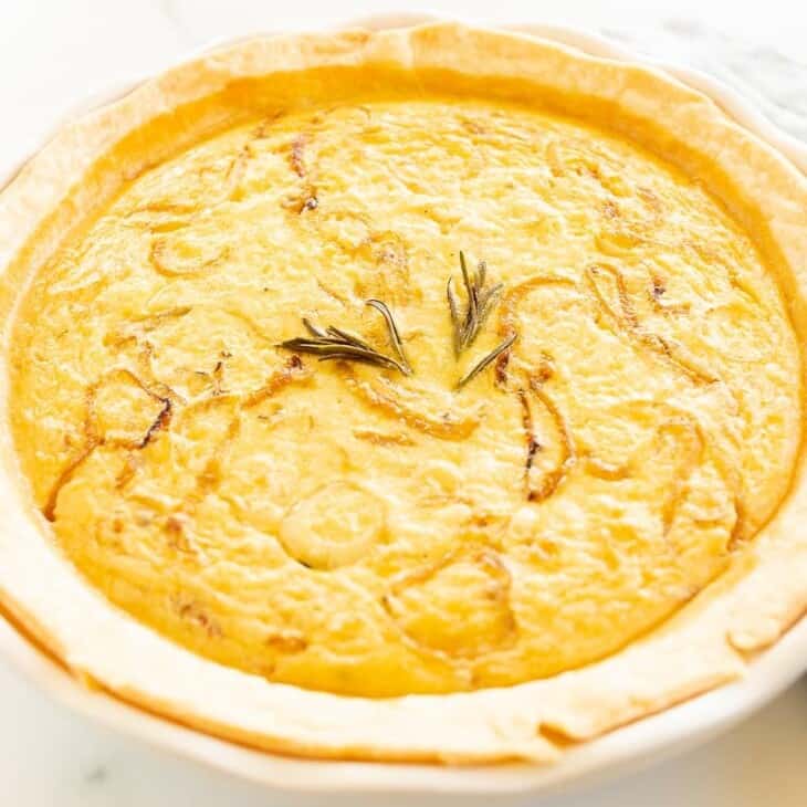 An onion quiche baked in a white pie pan on a marble surface.