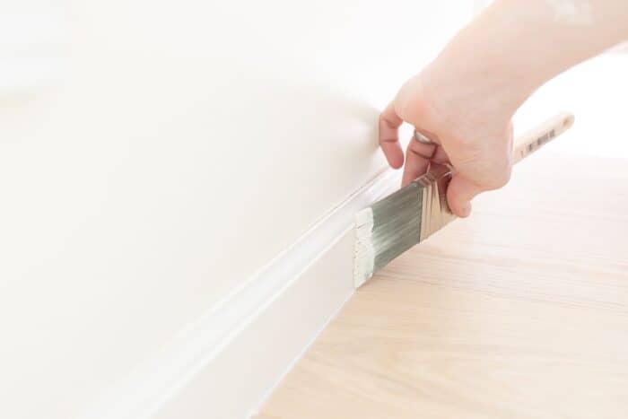 A hand painting trim white with a brush, next to a light wood floor.