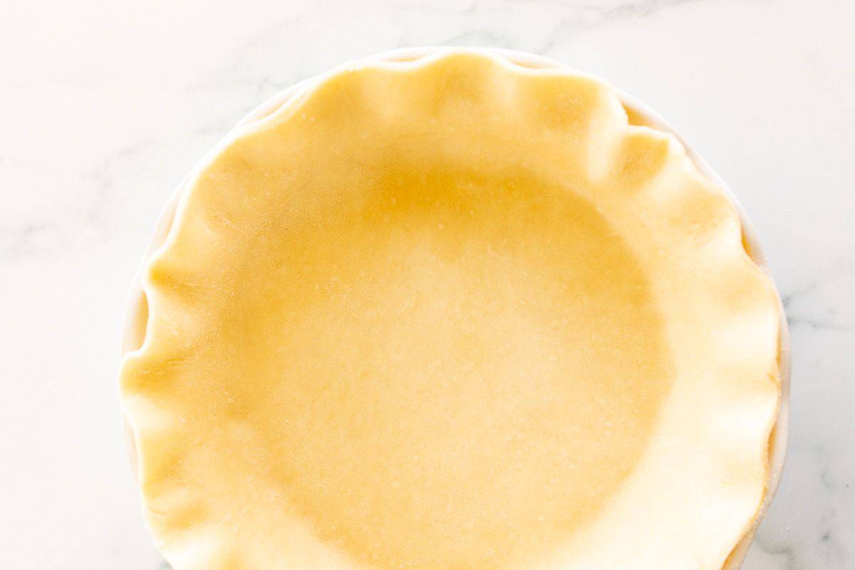 A homemade quiche crust in a fluted pie pan on a white marble surface.