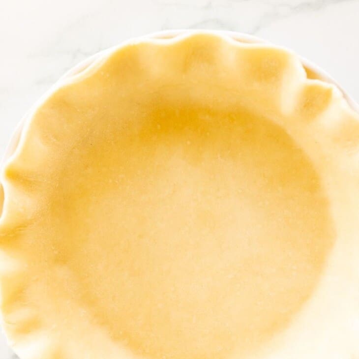 A homemade quiche crust in a fluted pie pan on a white marble surface.