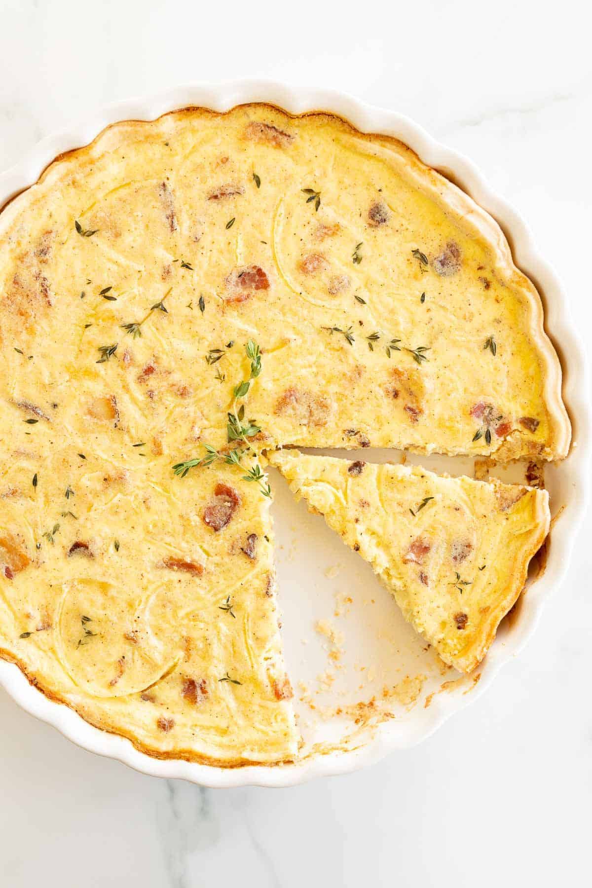 A homemade quiche in a white pie pan on a marble surface, one slice removed.