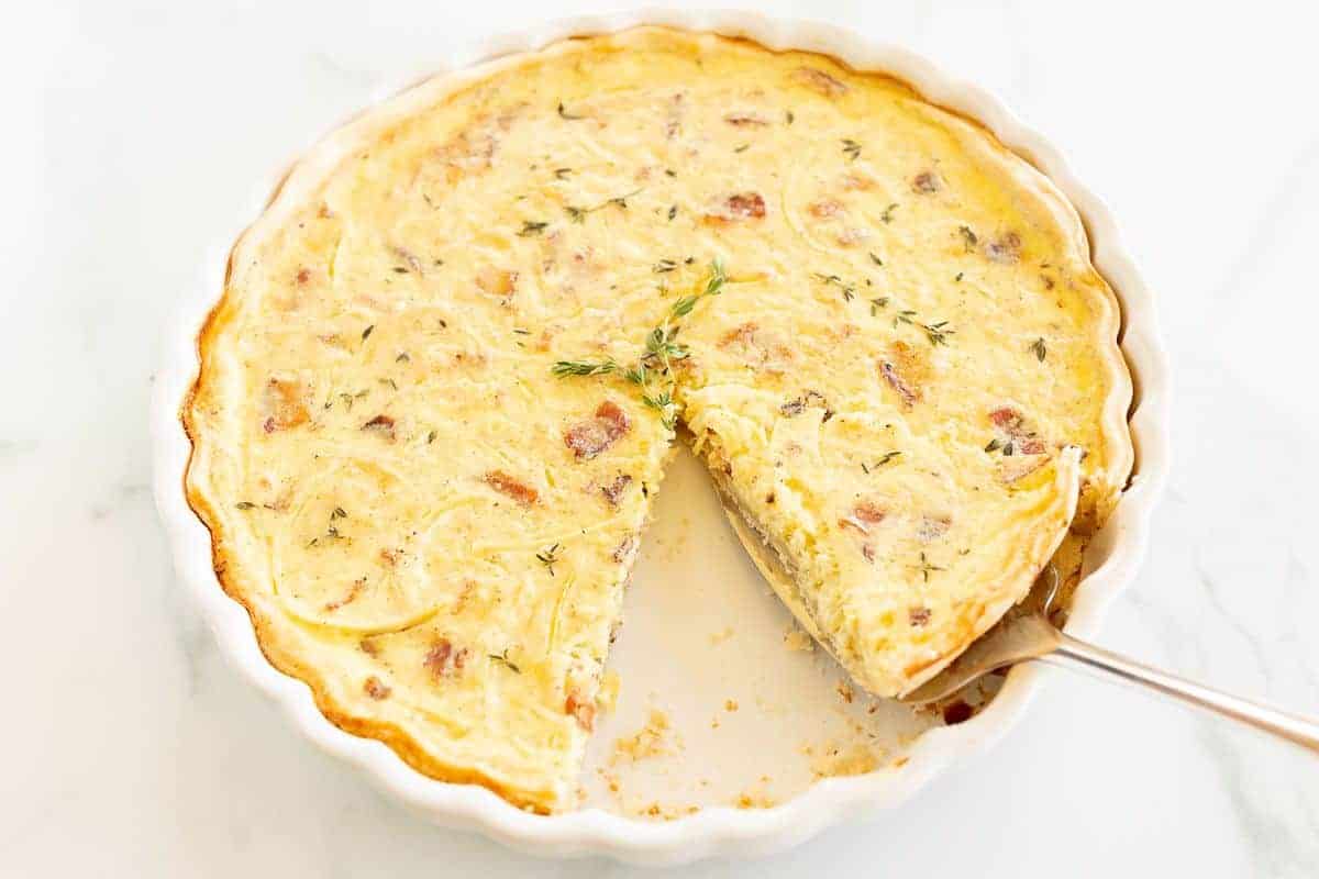 A whole quiche with a slice removed, pie server removing a second in a white pie dish.