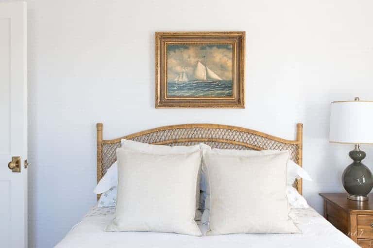 A bedroom with neutral bedding, a rattan bed, and sailboat art above headboard.