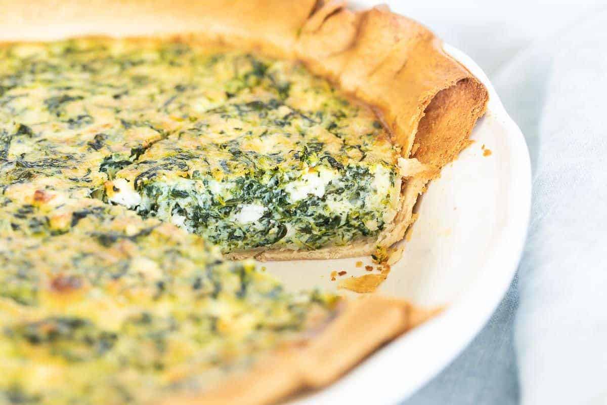 A spinach and feta quiche in a white pie pan, one slice removed.