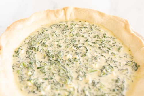 Spinach and feta quiche recipe, unbaked in a pie pan on a white surface.