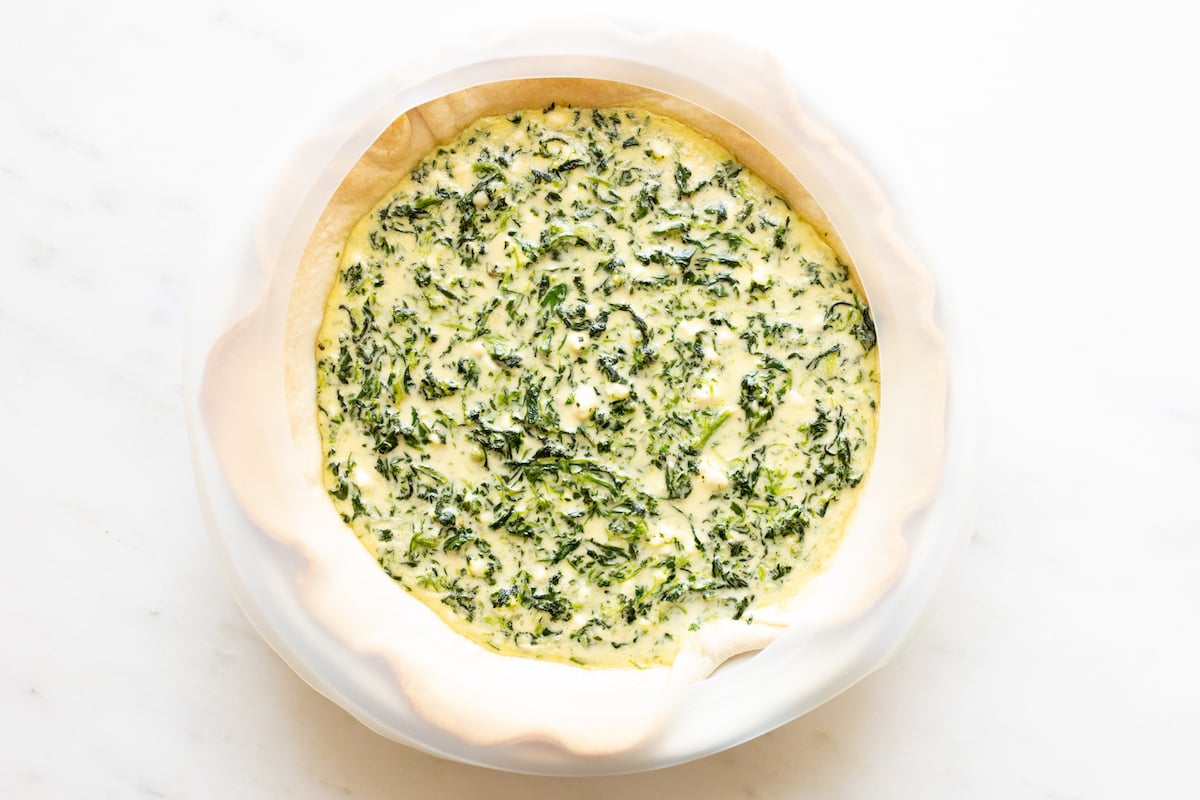Spinach and feta quiche recipe, unbaked in a pie pan on a white surface.