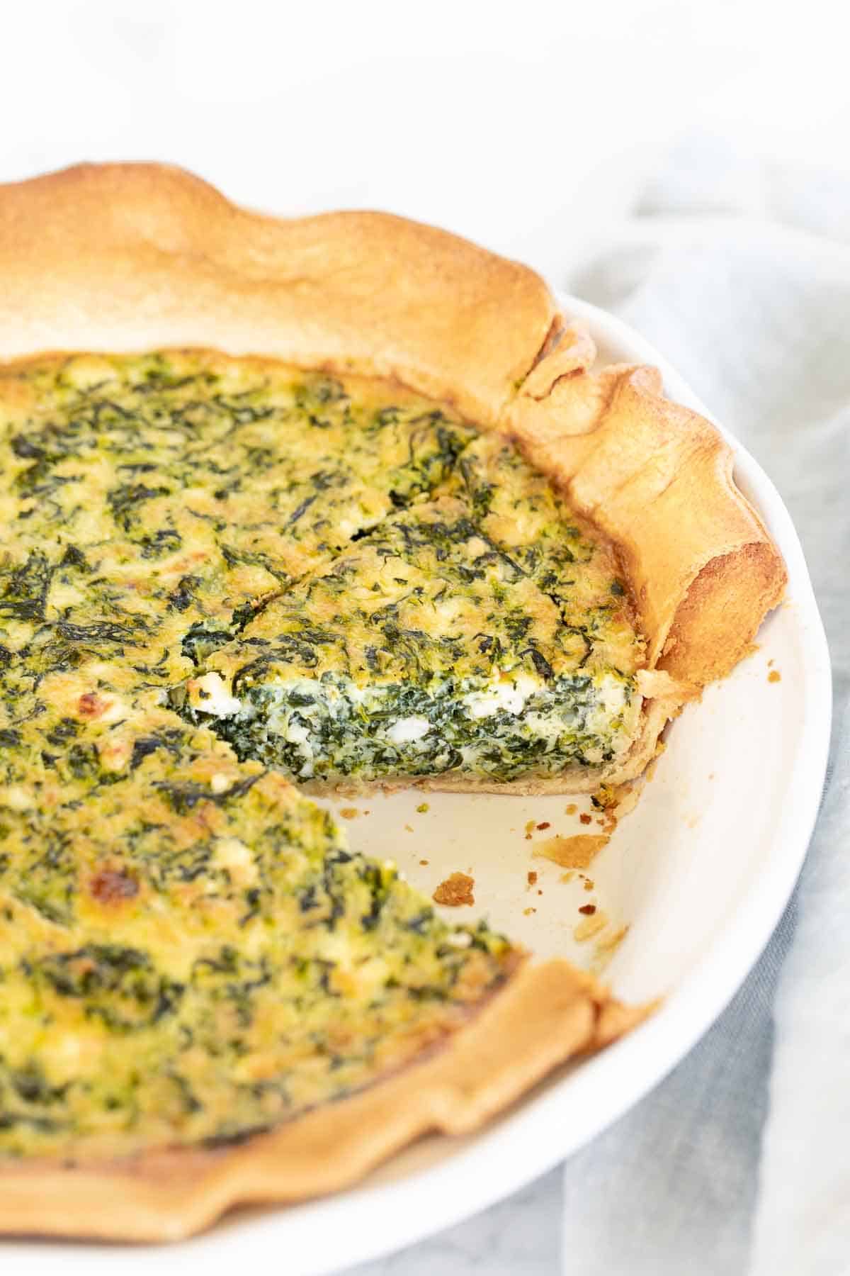 A spinach and feta quiche in a white pie pan, one slice removed.