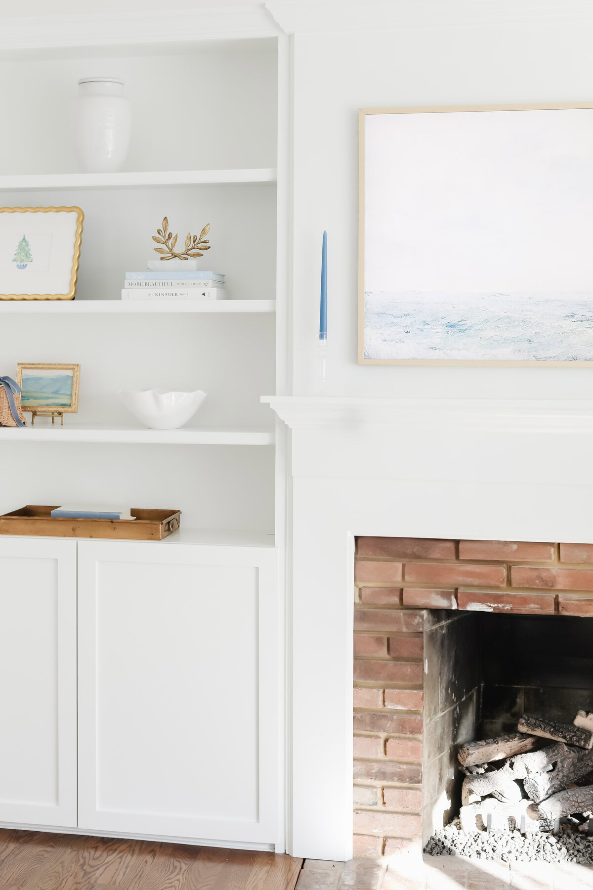 A white built-in bookshelf next to a fireplace with a brick interior; shelves hold books, framed art, a vase, and decor. A Samsung Frame TV and blue candle sit on the mantel, blending seamlessly with the painting and stylish ambiance.