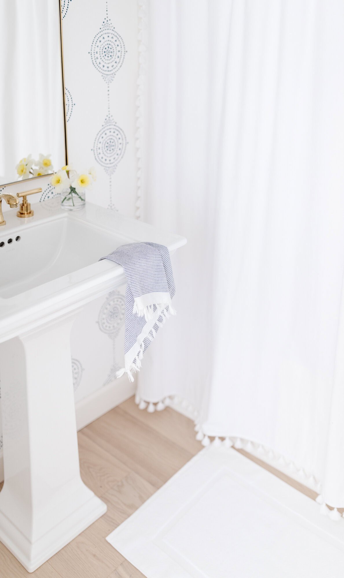 A white pedestal sink with a gold faucet, a blue towel, a vase of yellow flowers, and a white shower curtain in a bright bathroom with beautiful white oak floors.