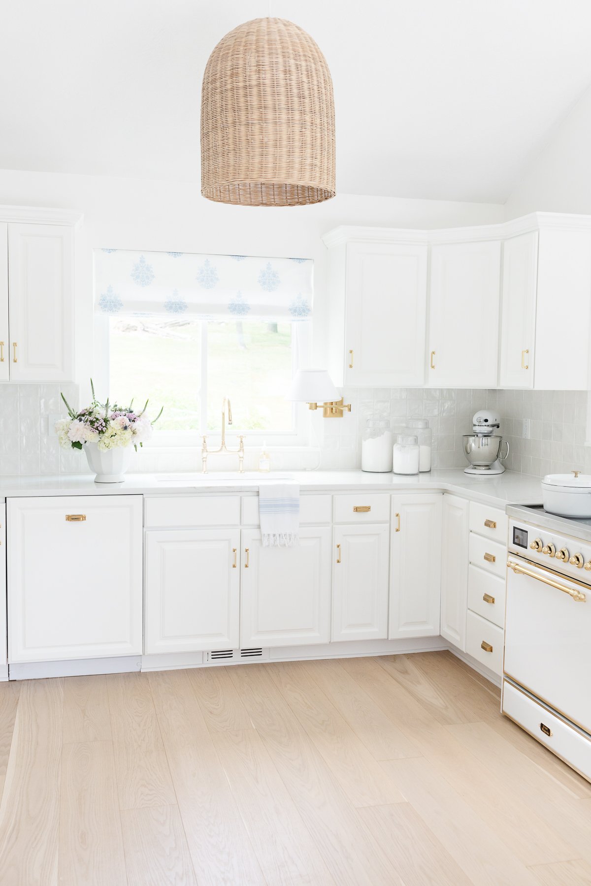 A bright, white kitchen features a wicker pendant light, white cabinets with brass handles, white oak floors, a bouquet of flowers on the counter, and a modern stove and mixer.