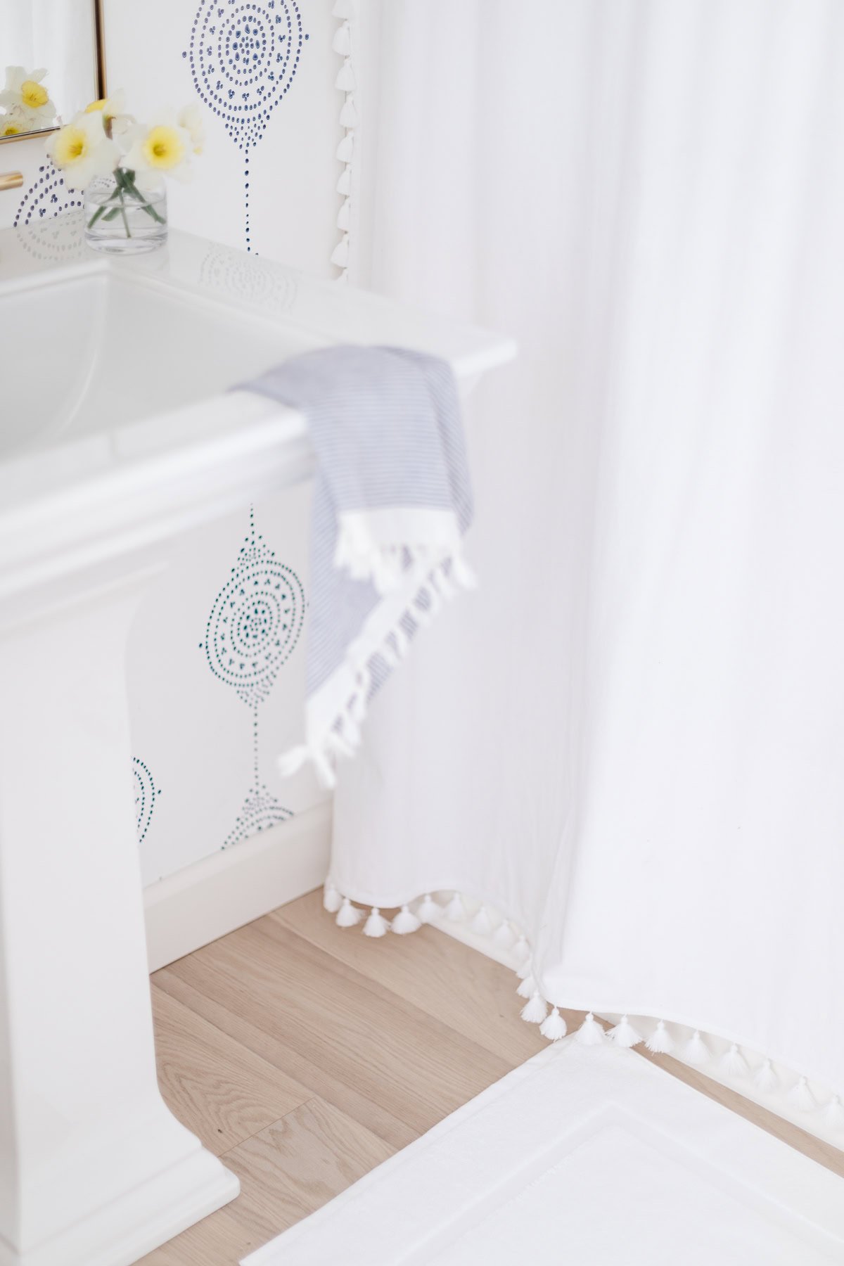 A bathroom with white oak floors, a white sink, blue and white striped hand towel, white shower curtain with tassels, and a small vase of yellow flowers on the counter.