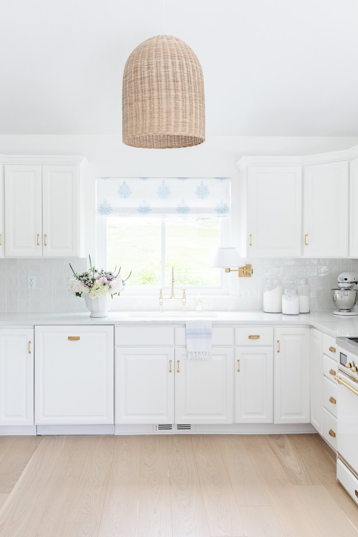 Bright white kitchen with gold hardware, white oak floors, a woven pendant light, a window with a patterned shade, and a vase of flowers on the counter.