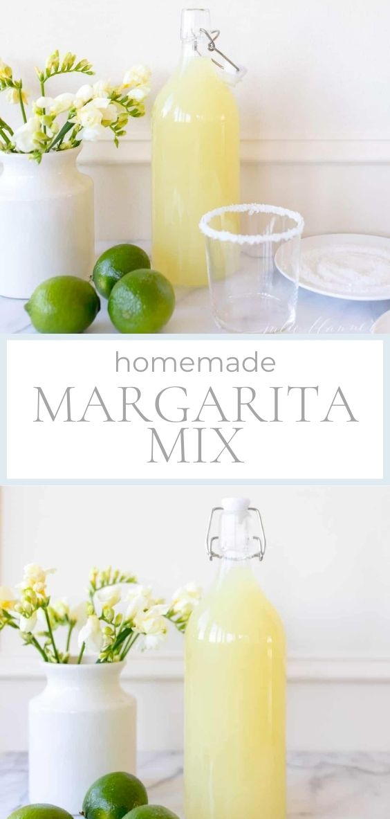 Homemade margarita mix is featured in a glass bottle on a marble counter with 3 limes a glass with a salt rim, and a vase of white and yellow flowers.