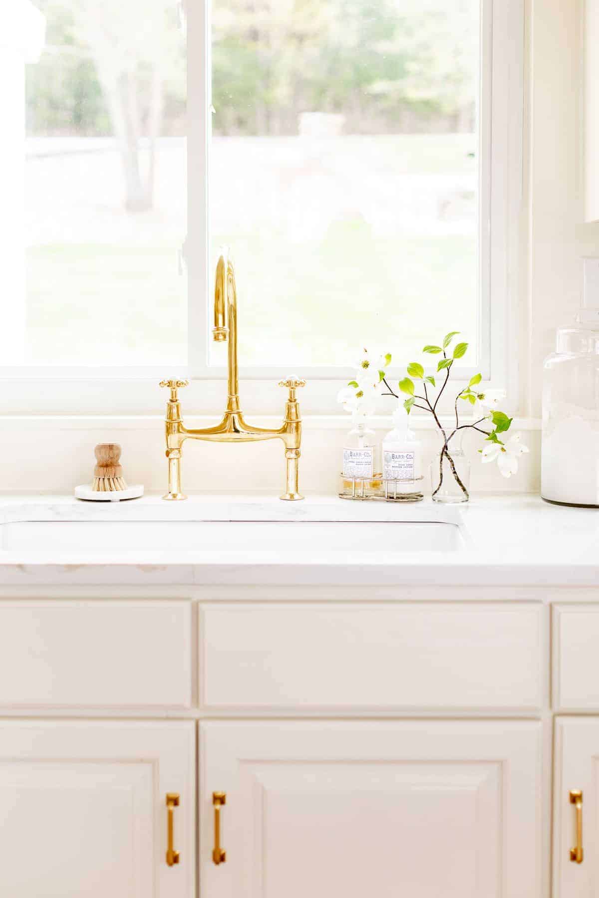 A white kitchen sink area with a brass bridge faucet, window over the sink and vase of flowers to the side.