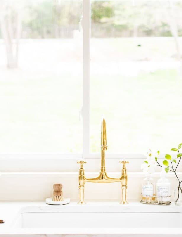 A white kitchen sink area with a brass bridge faucet, window over the sink and vase of flowers to the side.