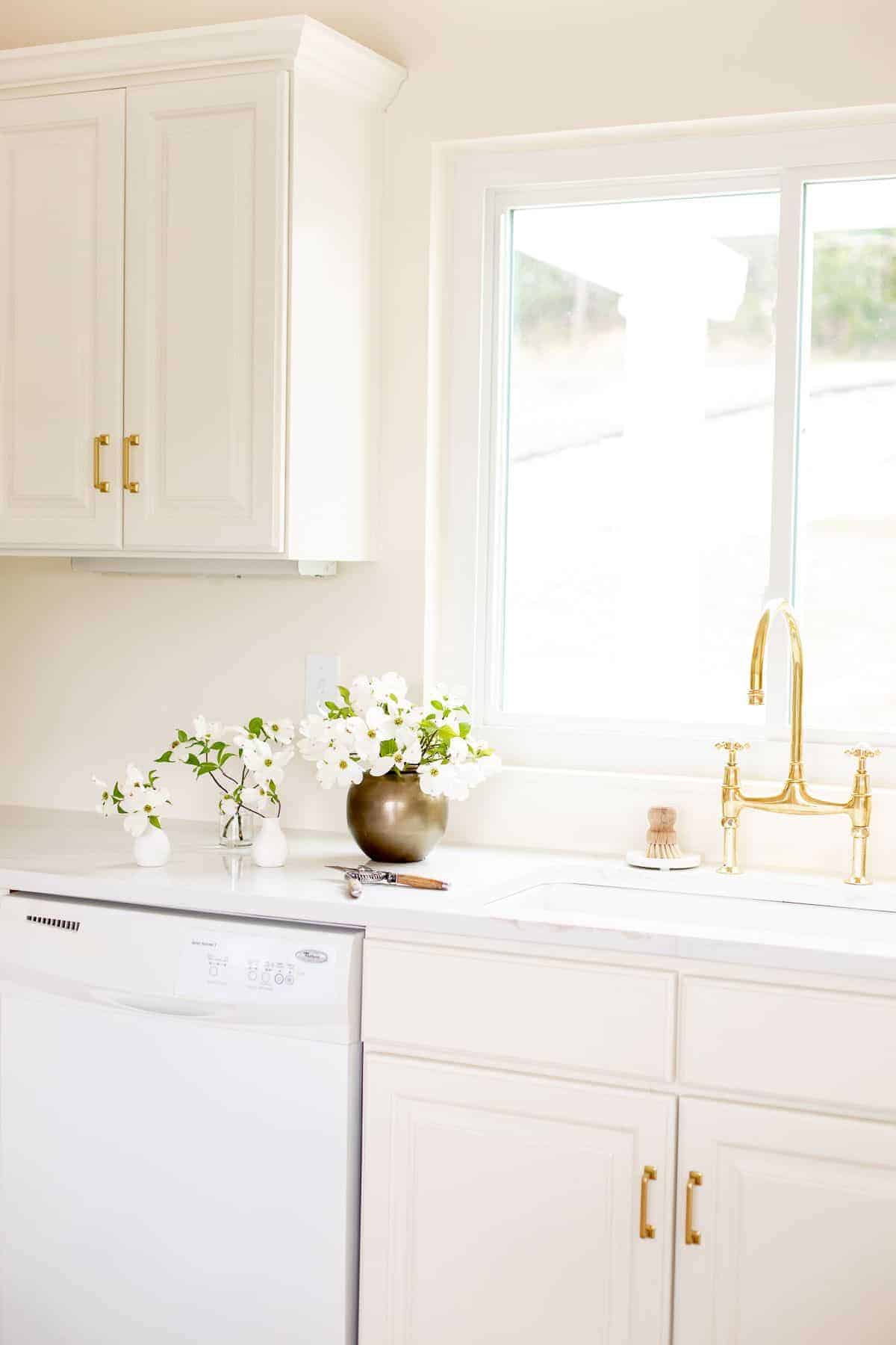 A white kitchen sink area with a brass bridge faucet, window over the sink and vase of flowers to the side.