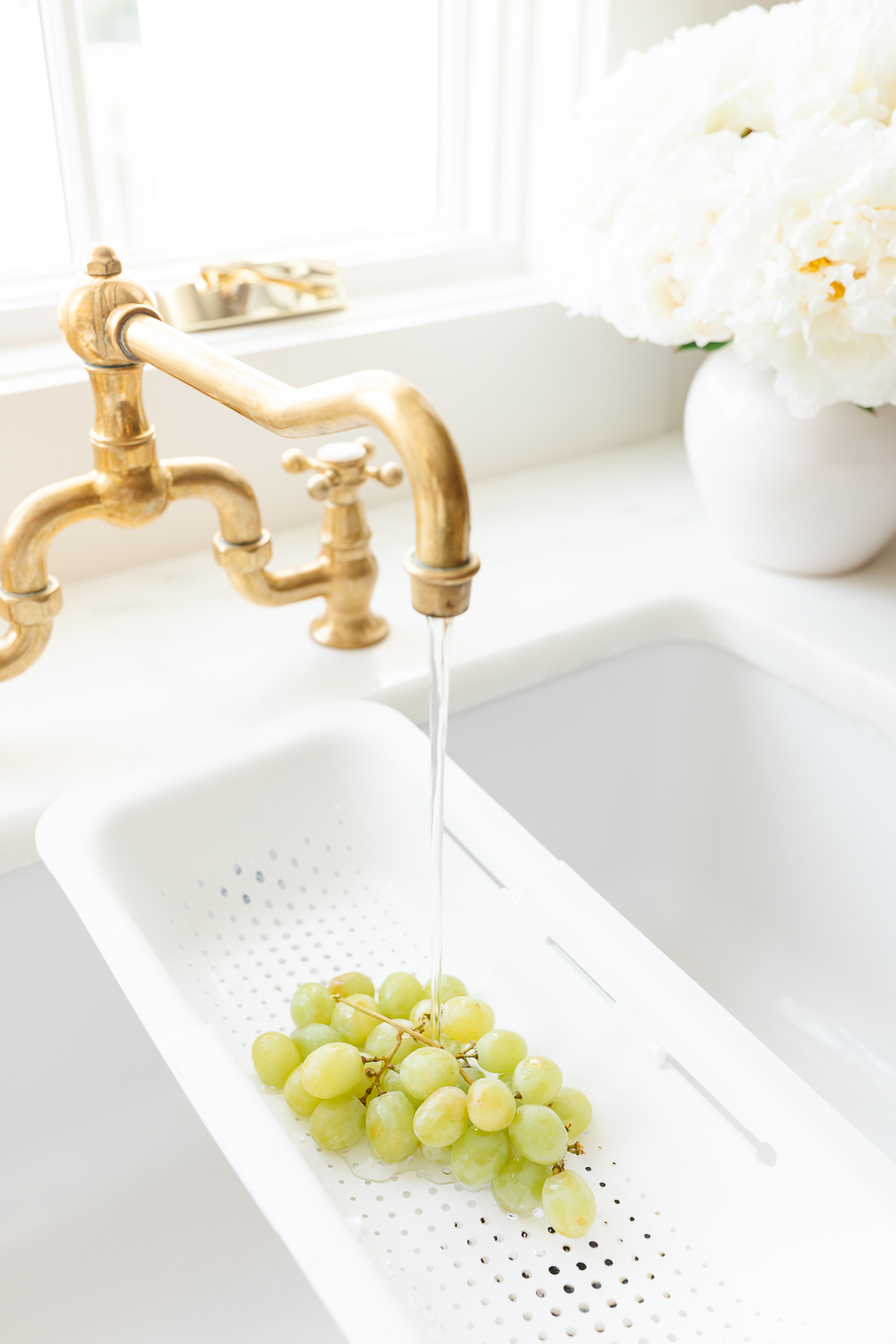 An unlacquered brass bridge kitchen faucet over a white farmhouse sink with grapes in a strainer. 