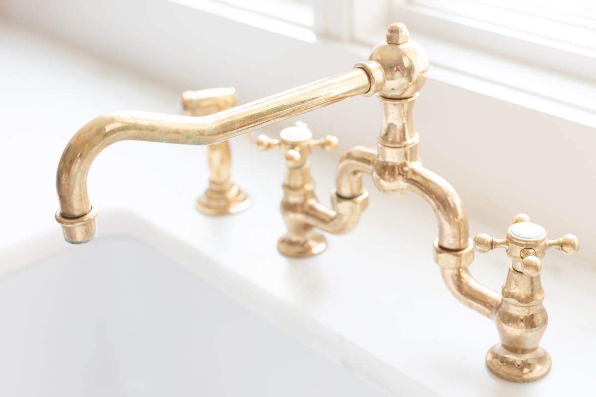 An unlacquered brass bridge kitchen faucet over a white farmhouse sink.