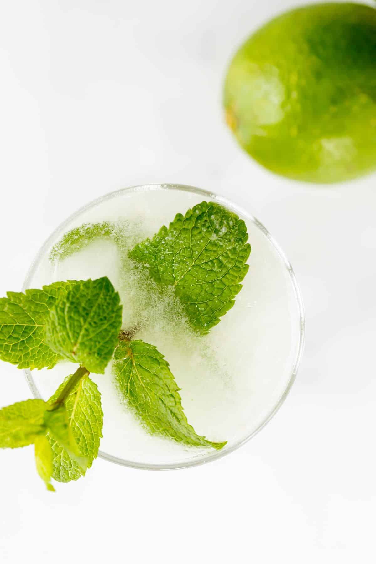 A white cocktail in a clear glass, garnished with fresh mint, lime in the background.