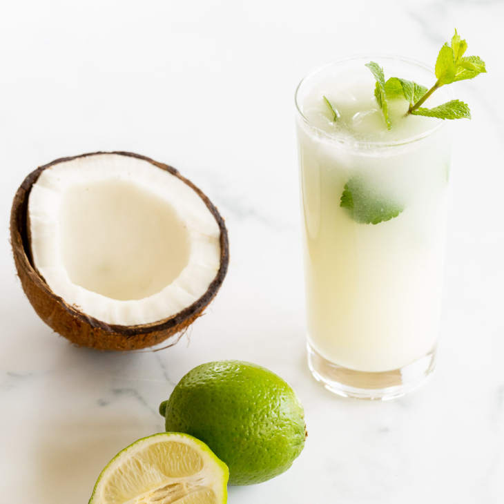 A coconut mojito garnished with mint on a marble surface. Limes and an open coconut to the side.