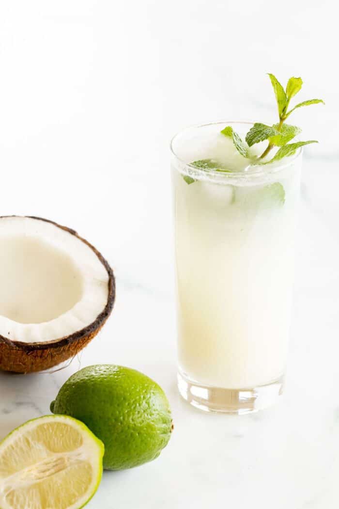 On a marble surface, a coconut mojito garnished with mint, half a coconut and a full lime near the glass.
