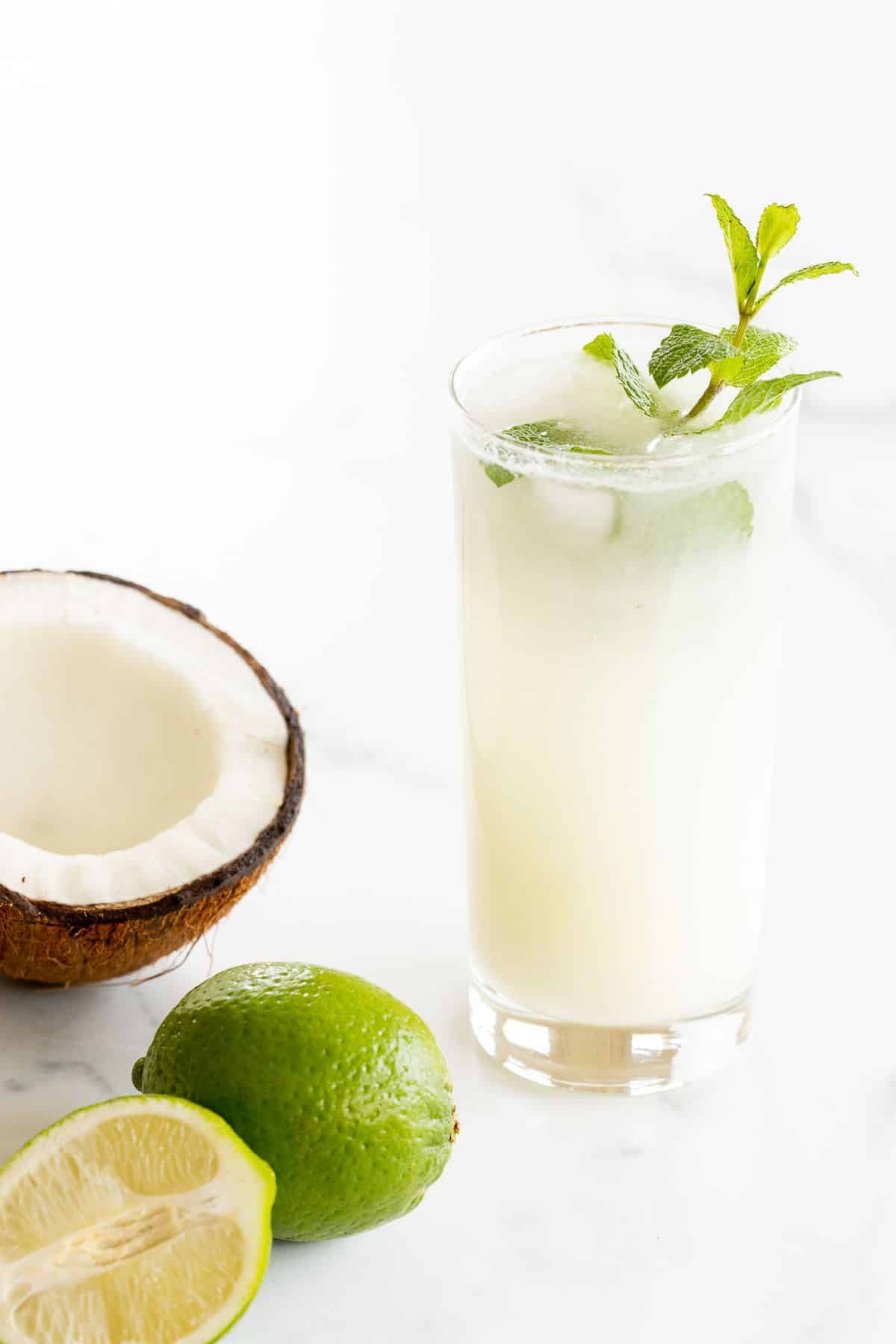 On a marble surface, a coconut mojito garnished with mint, half a coconut and a full lime near the glass.