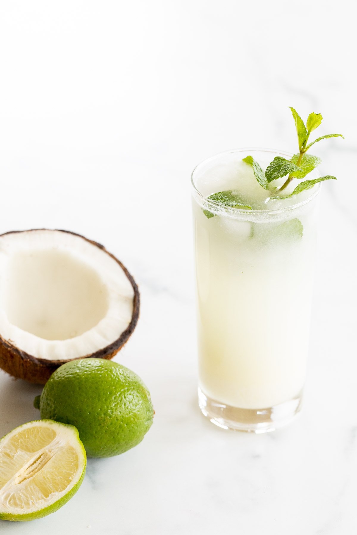 A coconut mojito garnished with mint on a marble surface. Limes and an open coconut to the side.