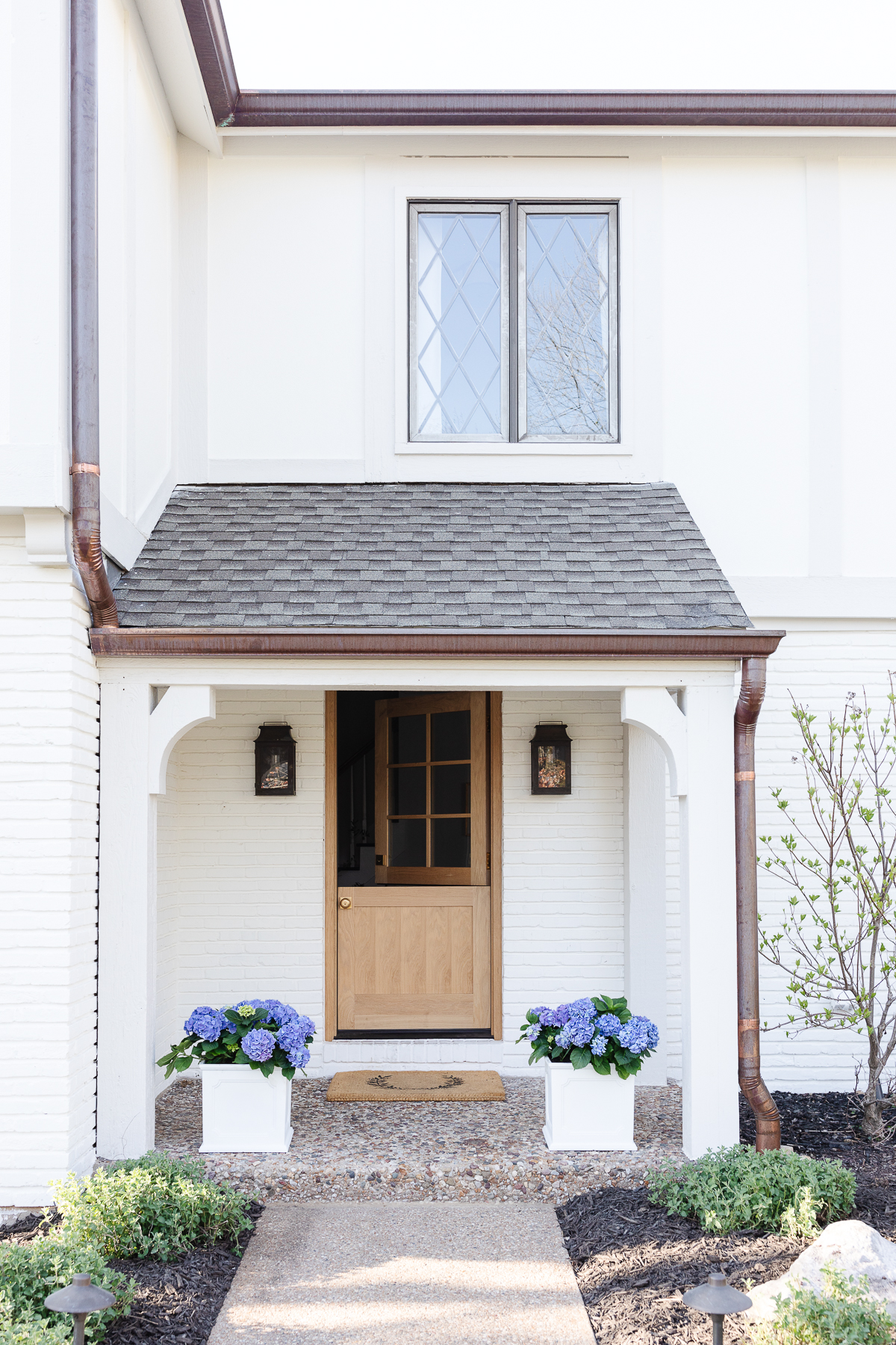 An entryway to a white brick home with copper lanterns on either side of the wooden Dutch door.