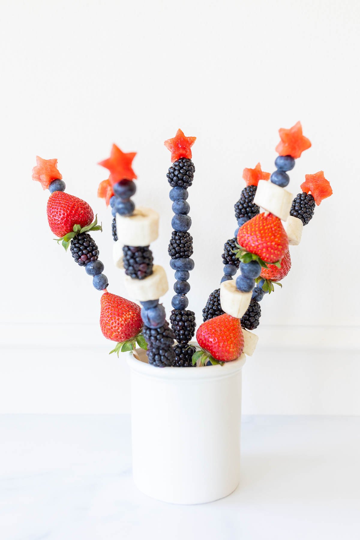 Red white and blue fruit skewers with a star on top in a white jar for display.