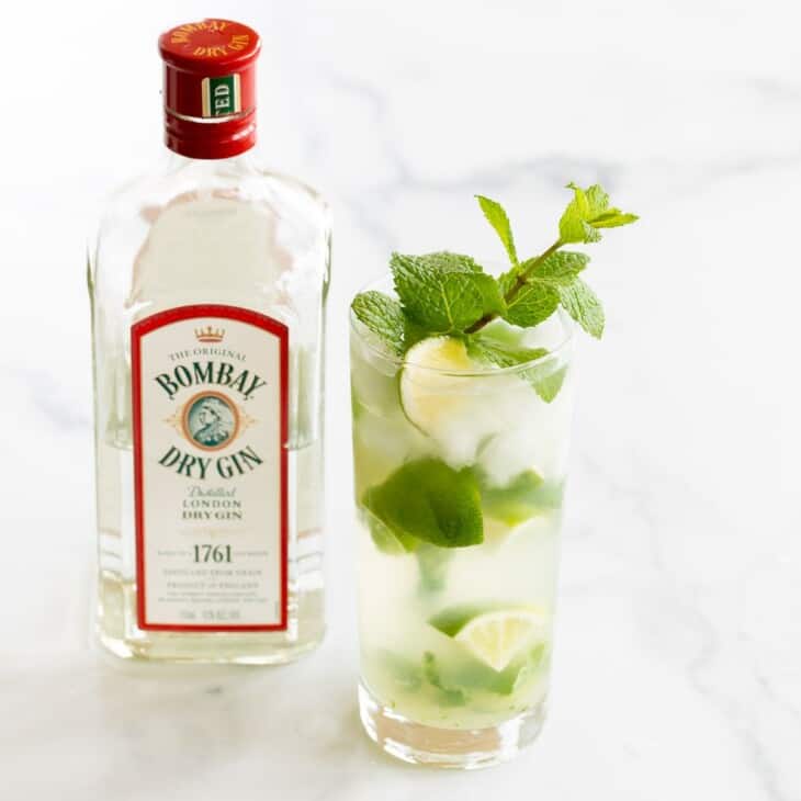A gin mojito in a clear glass, garnished with a wedge of lime and a sprig of mint. Gin bottle in background.