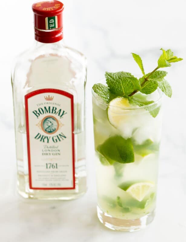 A gin mojito in a clear glass, garnished with a wedge of lime and a sprig of mint. Gin bottle in background.