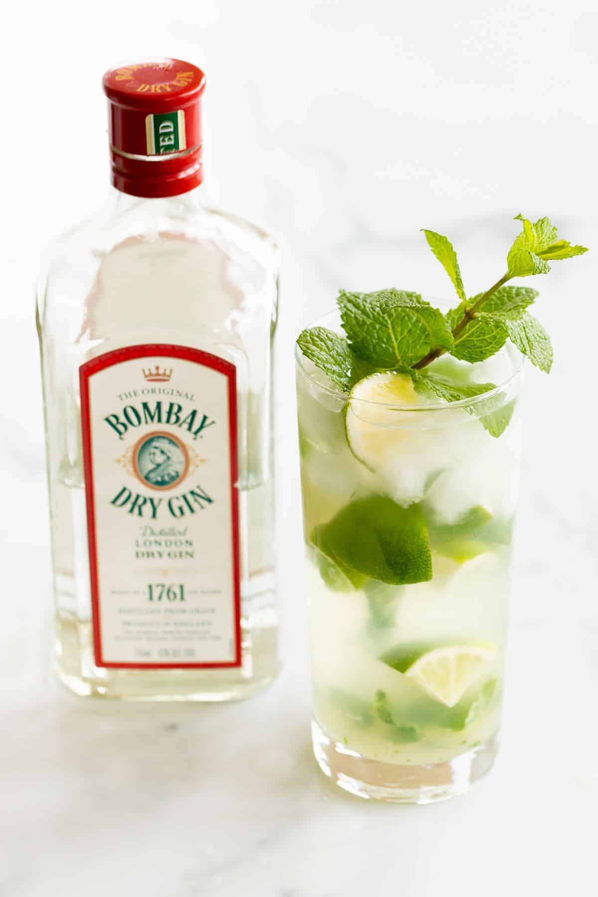 A gin mojito in a clear glass, garnished with a wedge of lime and a sprig of mint. Gin bottle in background.
