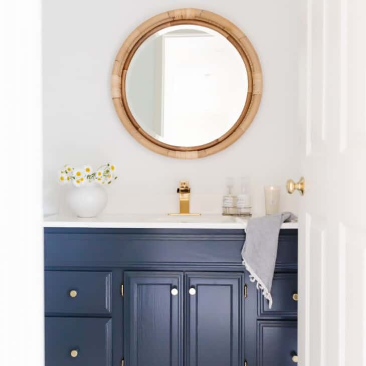 A white bathroom with a round rattan mirror and a dark blue vanity.