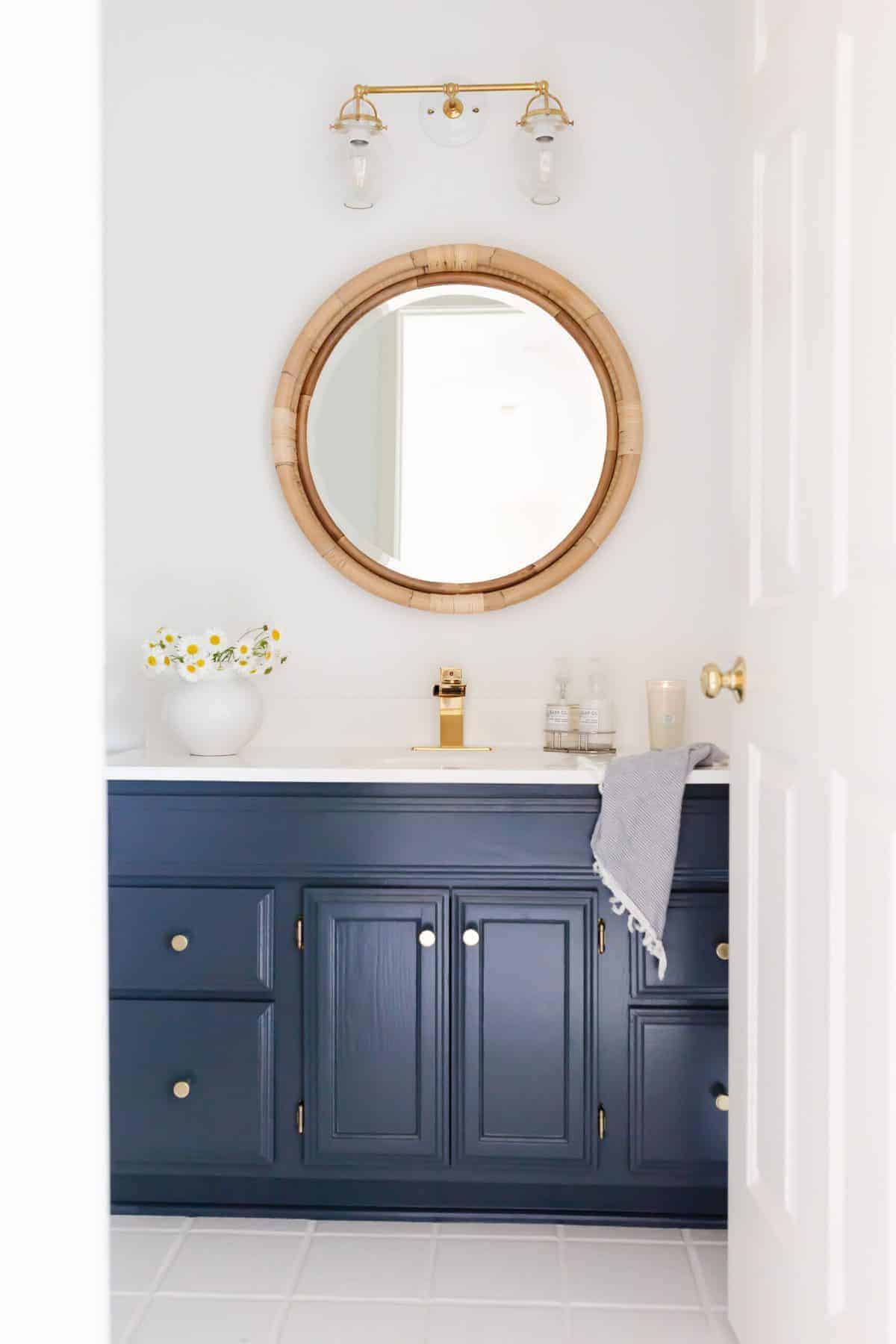 A white bathroom with a round rattan mirror and a dark blue vanity.