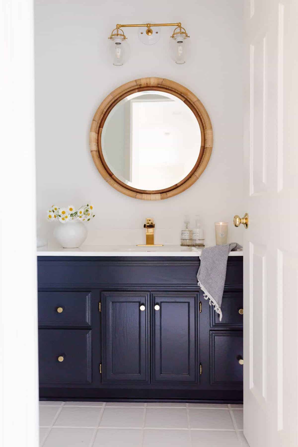 A white bathroom with a round rattan mirror and a dark blue vanity.