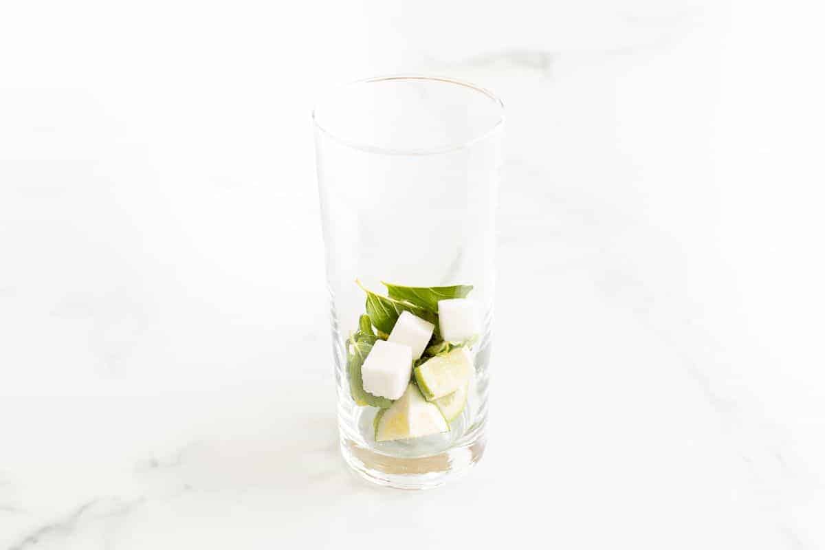A clear glass filled with sugar cubes, mint leaves and lime wedges on a marble surface.