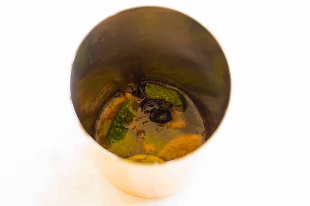 Looking inside a copper cocktail shaker, filled with muddled limes, mint and blueberries
