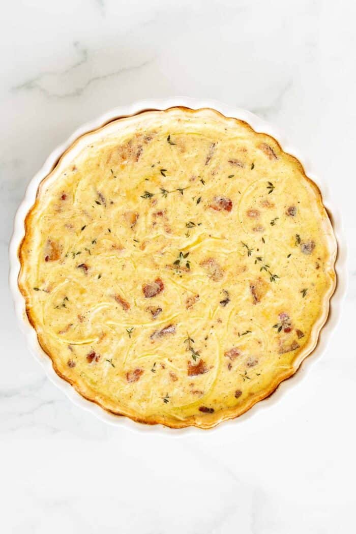 A quiche lorraine baked into a white tart pan, resting on a marble surface.