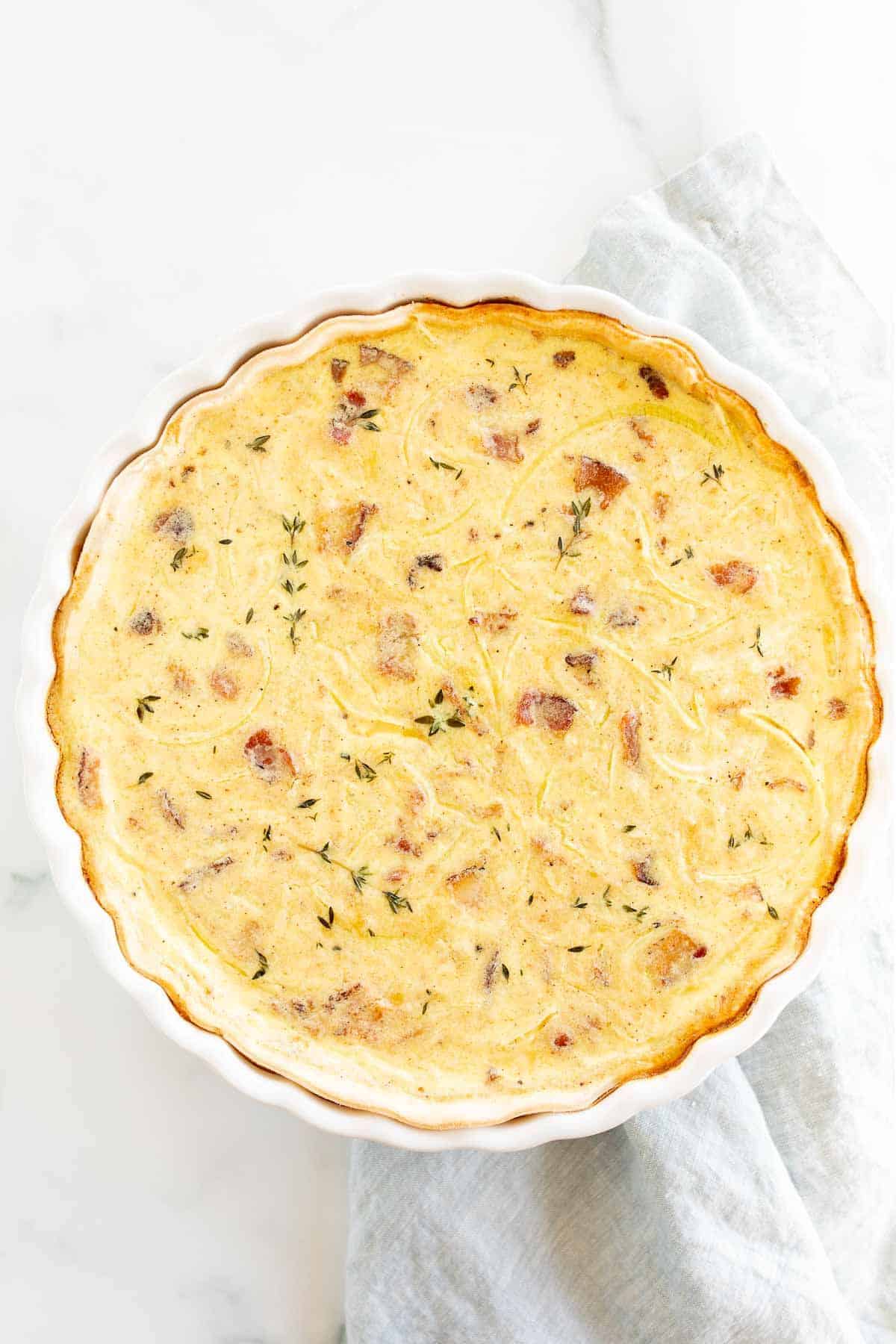 A quiche lorraine baked into a white tart pan, resting on a marble surface.