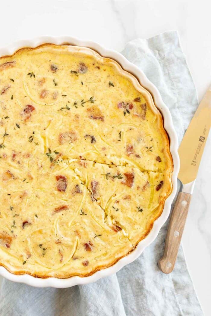 A quiche lorraine baked into a white tart pan, one slice cut out with knife to the side.