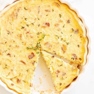 A quiche lorraine recipe in a white pan, one slice removed and another being cut away.