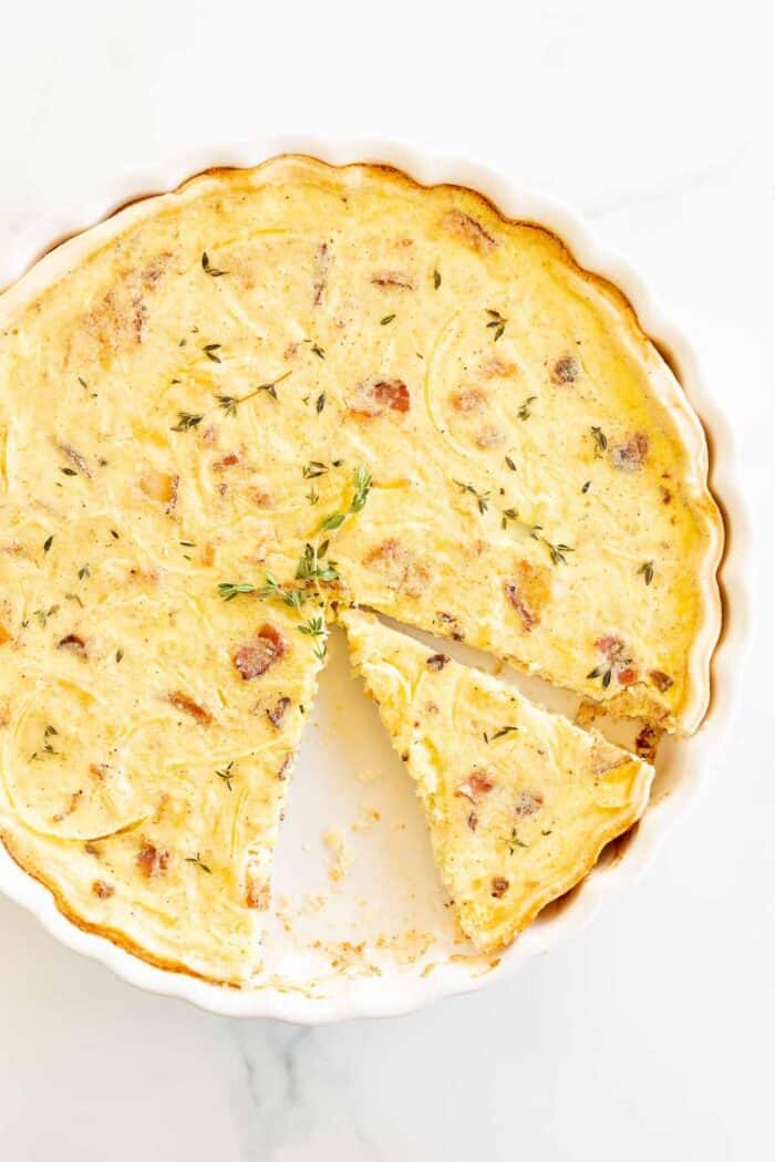 A quiche lorraine recipe in a white pan, one slice removed and another being cut away.
