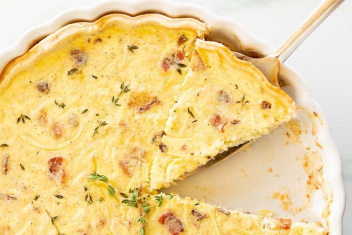 A quiche lorraine recipe in a white pan, one slice removed and another being cut away.