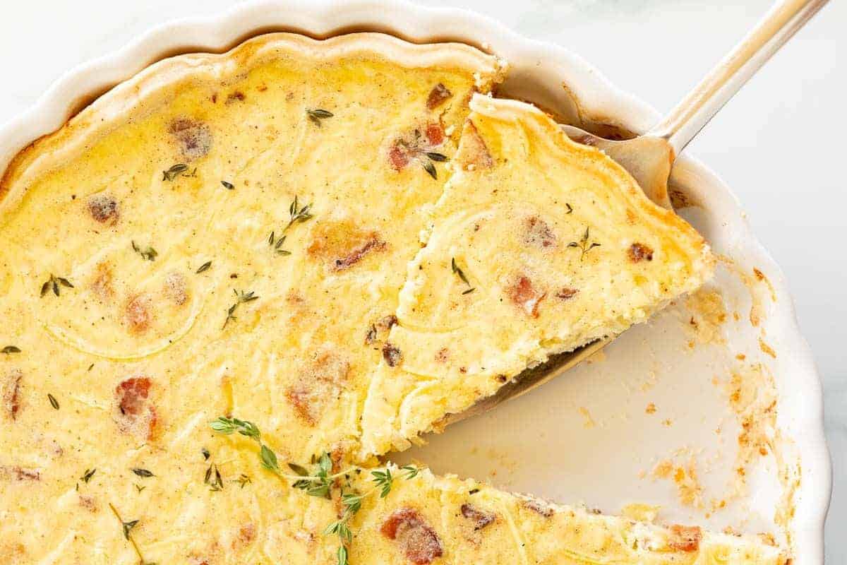 A quiche lorraine recipe in a white pan, one slice removed and another being cut away.