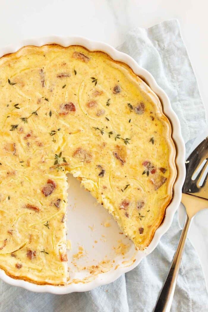 A quiche lorraine baked into a white tart pan, one slice cut out with serving utensil to the side.
